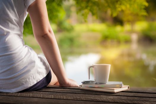 Young woman sitting and put down her hand beside coffee cup and notebooks on rustic wood bench with rural lake view in morning time on weekend