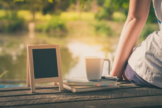 Young woman sitting and put down her hand beside coffee cup, notebooks and small A-frame blackboard on rustic wood bench in weekend with morning scene and vintage filter effect