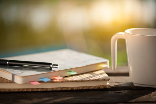 Pen, notebooks, and coffee cup on rustic wood table at outdoor area with morning scene in summer time