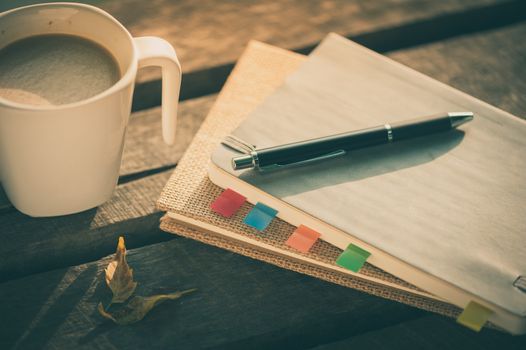 Pen, notebooks, and coffee cup on rustic wood table at outdoor area in morning time on summer with vintage filter effect