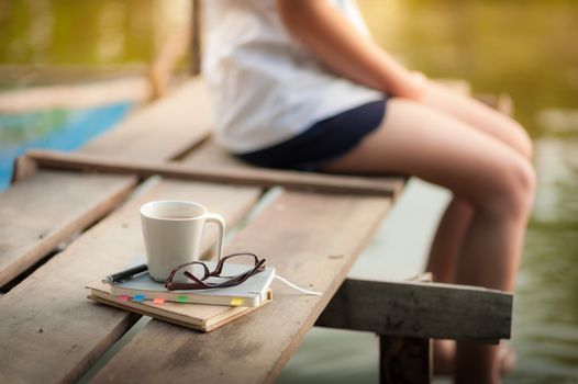 Notebooks, pen, glasses, and coffee cup are putting down on the wood waterside while young woman sitting beside them in morning time on weekend