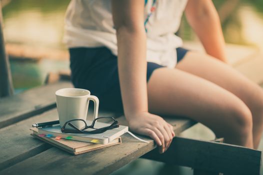Notebooks, pen, glasses, and coffee cup are putting down on the wood waterside while young woman sitting beside them in morning time on weekend with vintage filter effect