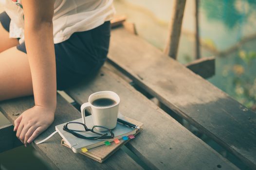 Notebooks, pen, glasses, and coffee cup are putting down on the wood waterside while young woman sitting beside them in morning time on weekend with vintage filter effect
