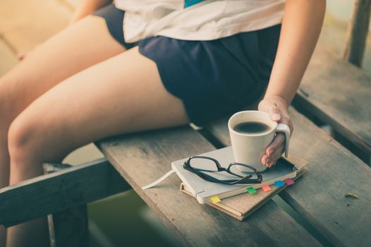 Notebooks, pen, glasses, and coffee cup are putting down on the wood waterside with young woman left hand holding coffee cup in morning time on weekend with vintage filter effect