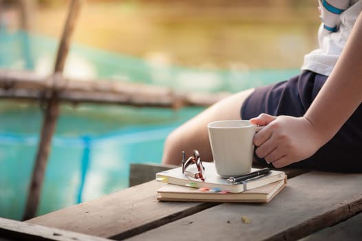 Young woman left hand holding coffee cup above notebokks on wood waterside in morning time on weekend