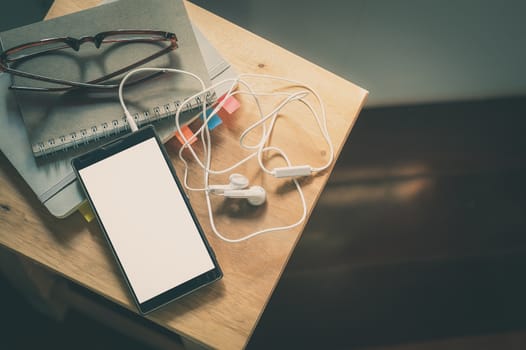 Smartphone with small headphones, glasses, and notebokks on wood chair near to window in morning time on weekend with vintage filter effect
