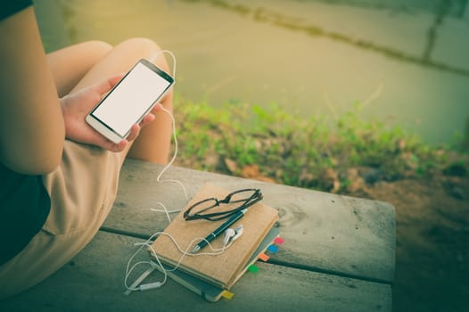 Young woman holding smartphone with small headphones by left hand while sitting on wood bench in park in morning time on weekend with vintage filter effect