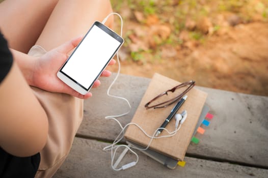 Young woman holding smartphone with small headphones by left hand while sitting on wood bench in park in morning time on weekend