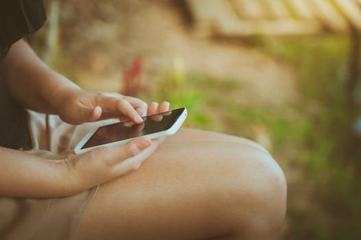 Young woman touching on smartphone screen while sitting on wood bench in park in morning time on weekend with vintage filter effect