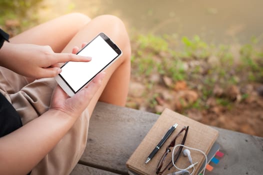 Young woman touching on smartphone blank screen while sitting on wood bench in park in morning time on weekend