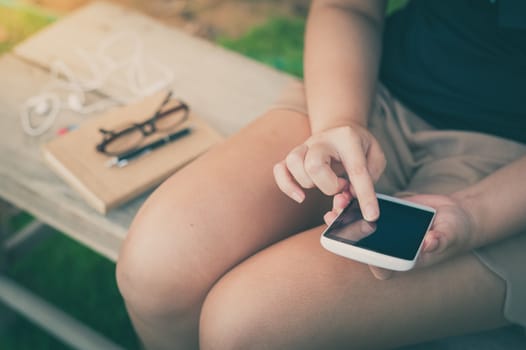 Young woman touching on smartphone screen while sitting on wood bench in park in morning time on weekend with vintage filter effect