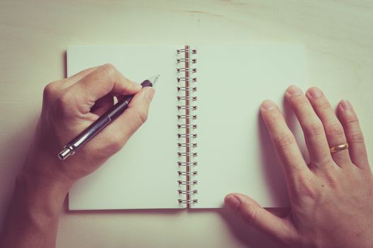 Male left hand writing on opened notebook with blank area for text or message and pen on wood table with vintage filter effect