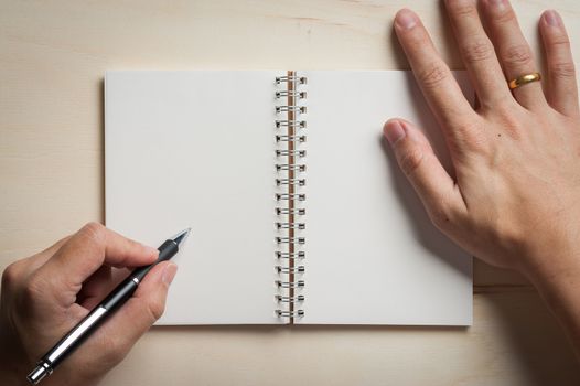 Male left hand writing on opened notebook with blank area for text or message and pen on wood table in morning time
