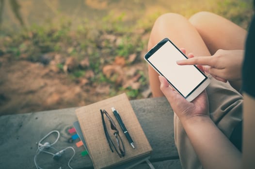 Young woman touching on smartphone blank screen while sitting on wood bench in park in morning time on weekend with vintage filter effect