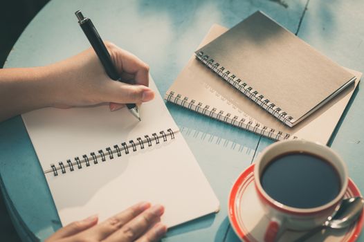 Woman left hand writing journal on small notebook in outdoor area at cafe with morning scene and vintage filter effect