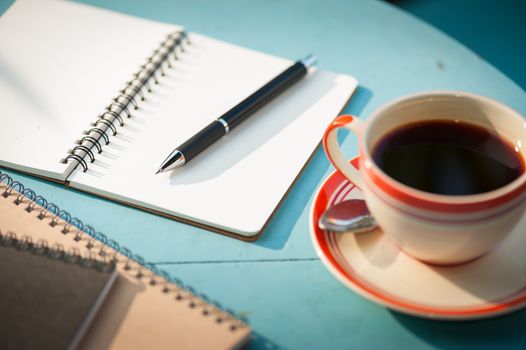 Small notebook and pen beside coffee cup on blue wood table at outdoor area in cafe with morning scene