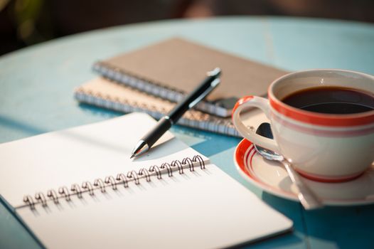 Small notebook and pen beside coffee cup on blue wood table at outdoor area in cafe with morning scene