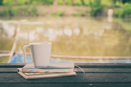 Coffee cup and notebooks on rustic wood table at outdoor area with rural lake view on blurry background in morning time with vintage filter effect