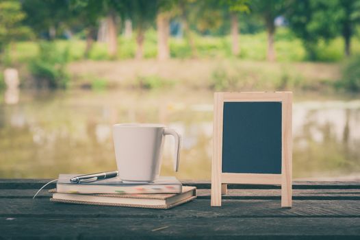 Small A-frame blackboard with blank area for text or message, notebooks and coffee cup on rustic wood table with rural lake view in blurry background at morning time with vintage filter effect