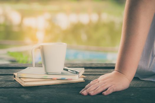 Woman hand put down beside coffee cup and notebooks on rustic wood bench with rural lake view in morning time on weekend with vintage filter effect