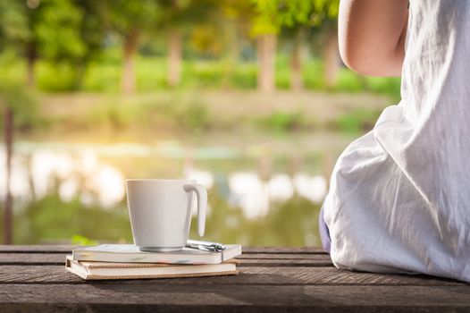 Young woman sitting beside coffee cup and notebooks on rustic wood bench with rural lake view in morning time on weekend