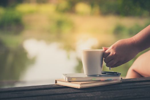Woman left hand holding coffee cup beside notebooks on rustic wood bench in morning time on weekend with vintage filter effect