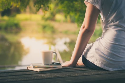 Young woman sitting and put down her hand beside coffee cup and notebooks on rustic wood bench with rural lake view in morning time on weekend