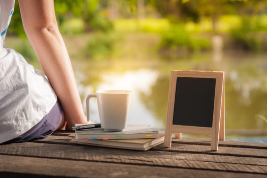 Young woman sitting and put down her hand beside coffee cup, notebooks and small A-frame blackboard on rustic wood bench in weekend with morning scene