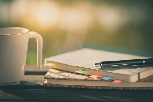 Pen, notebooks, and coffee cup on rustic wood table at outdoor area in morning time in summer with vintage filter effect
