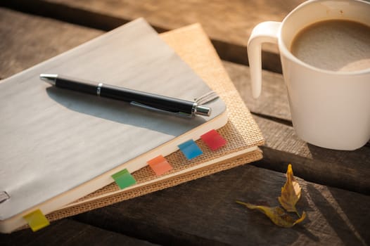 Pen, notebooks, and coffee cup on rustic wood table at outdoor area with morning scene in summer time