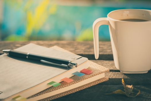 Pen, notebooks, and coffee cup on rustic wood table at outdoor area with morning scene in summer time with vintage filter effect