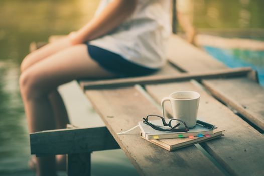 Notebooks, pen, glasses, and coffee cup are putting down on the wood waterside while young woman sitting beside them in morning time on weekend with vintage filter effect