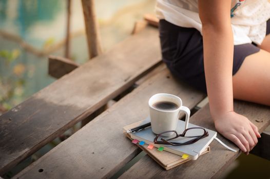 Notebooks, pen, glasses, and coffee cup are putting down on the wood waterside while young woman sitting beside them in morning time on weekend