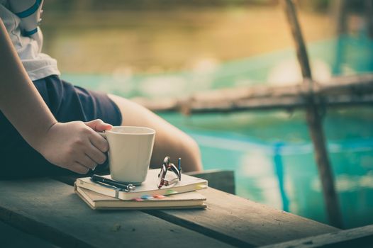 Young woman right hand holding coffee cup above notebokks on wood waterside in morning time on weekend