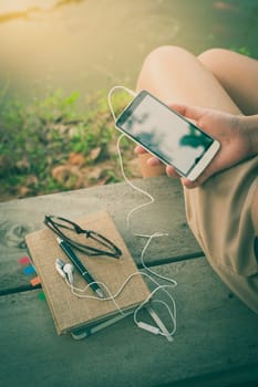 Young woman  holding smartphone with small headphones by right hand while sitting on wood bench in park in morning time on weekend with vintage filter effect