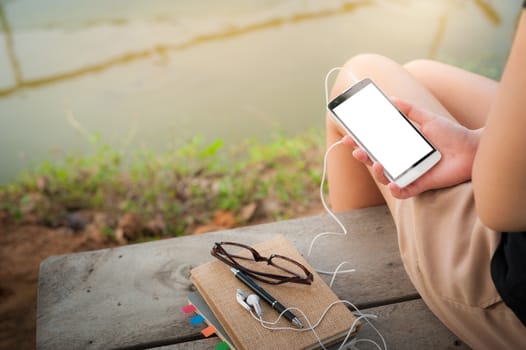 Young woman holding smartphone with small headphones by right hand while sitting on wood bench in park in morning time on weekend