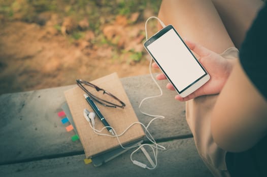 Young woman holding smartphone with small headphones by right hand while sitting on wood bench in park in morning time on weekend with vintage filter effect