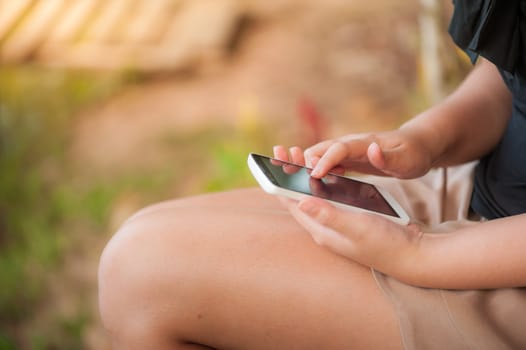 Young woman touching on smartphone screen while sitting on wood bench in park in morning time on weekend