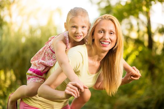 Beautiful mother giving her cute little girl piggyback and smiling in the park