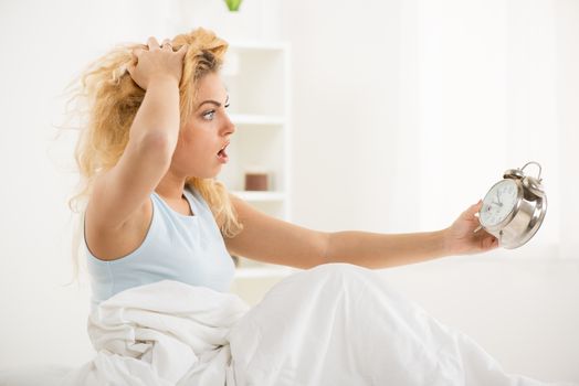 Cute young woman awakening in the morning. She is oversleep and looking with alarm clock.