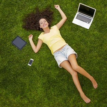 Beautiful and happy young woman lying on the grass surrounded by technology devices