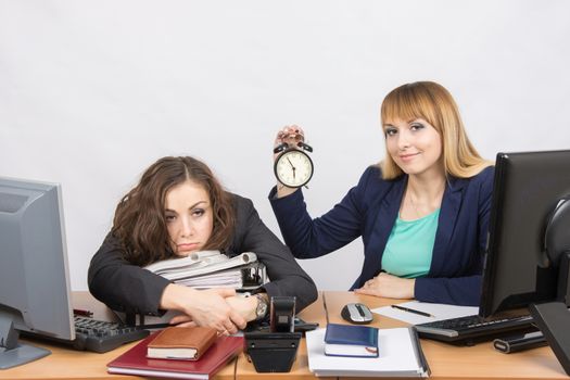 Two girls in the office at the end of the day, one with a smile, holding a clock, another weary lies on folders