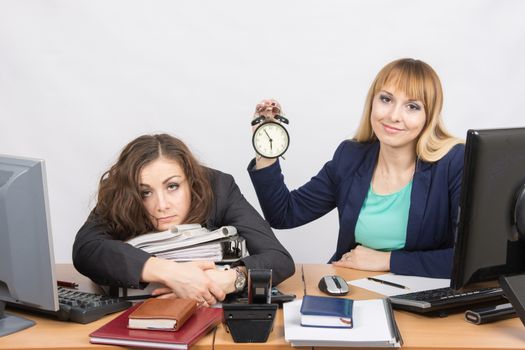 The girl in the office with a smile shows the time on the clock next to the exhausted colleague