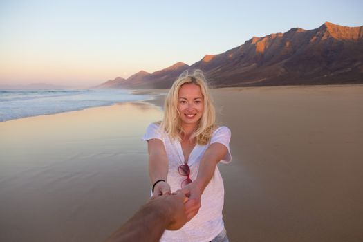 Young romantic couple, holding hands, having fun on perfect deserted beach at sunset. Shot from boyfrieds perspective. Guy looking at her beautiful carefree girlfriend.