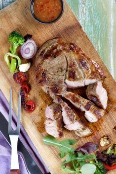 Delicious Roasted Pork Neck with Grilled Vegetables, Greens and Hot Sauce Sliced on Wooden Cutting Board with Meat Fork. Top View