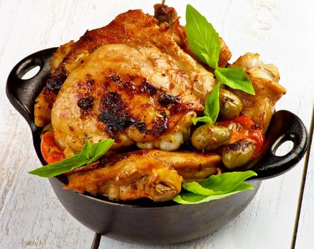 Delicious Crispy Roasted Chicken Thighs with Tomatoes, Green Olives and Basil in Black Fry Pan closeup on White Wooden background