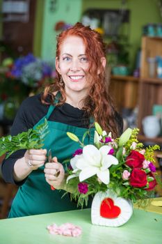 Pretty woman in green apron creating flower arrangement at table in florist shop