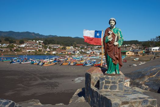 Colourfully painted statue of Saint Peter along side the flag of Chile on a rocky promontory sheltering the beach used by the fishing fleet in the small fishing village of Curanipe in the Maule Region of Chile. Fishing boats beyond.