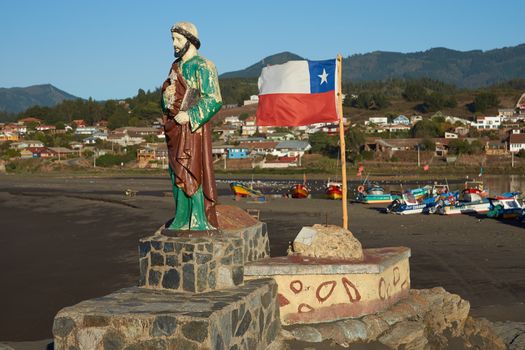 Colourfully painted statue of Saint Peter along side the flag of Chile on a rocky promontory sheltering the beach used by the fishing fleet in the small fishing village of Curanipe in the Maule Region of Chile. Fishing boats beyond.