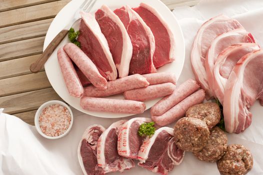Assorted raw meat cuts with salt including pork cutlets, steak, lamb chops, sausage and meatballs on a wooden table